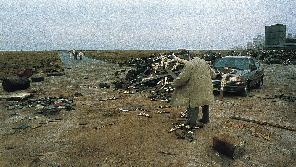 A wide plain of land with a long road heading away from the image centre under overcast sky. A man in a brown coat looks out at someone walking down the road towards him. He is next to an old cadilliac car, suitcases and a refuse pile of large, wooden crucifixes.
