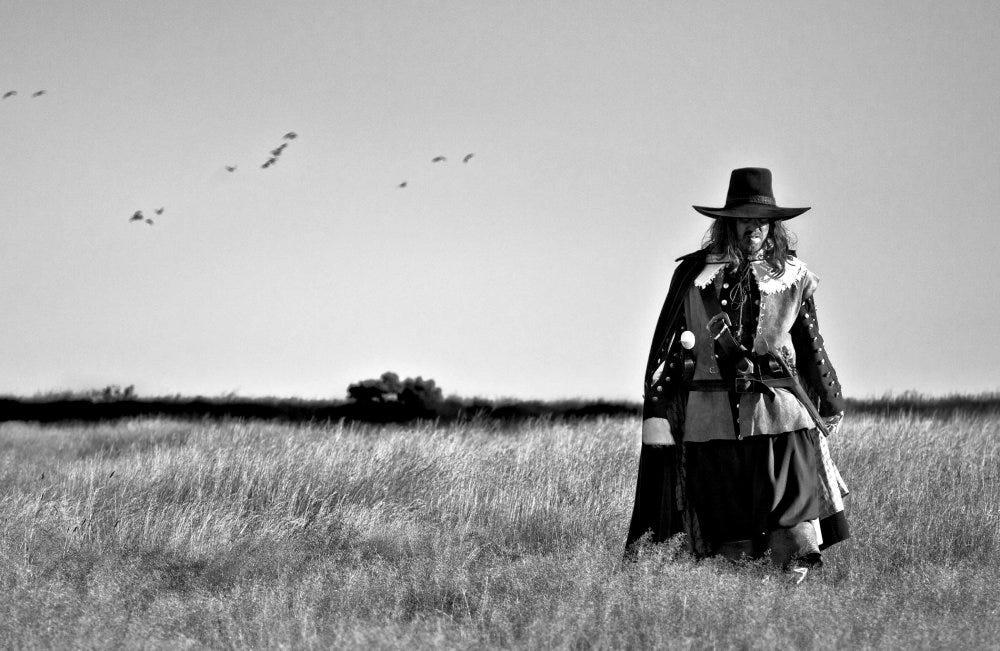 A black and white still of the film ‘a field in England’ showing a 16th century English soldier with a wide brimmed hat and cloak walking through a wheat field with birds overhead. His face and eyes can’t be seen clearly.