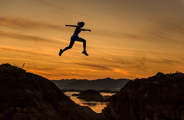 A woman leaping between two hills over a large river. a dark, orange sunset is in the background.
