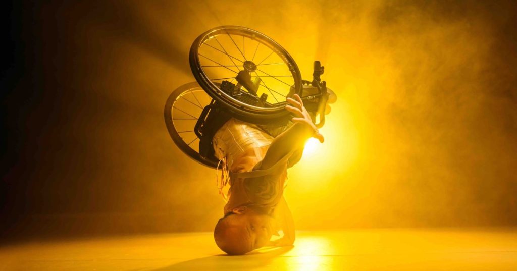 A performer on stage, they are balancing on their head with their wheelchair in the air, behind them is a bright orange spotlight.