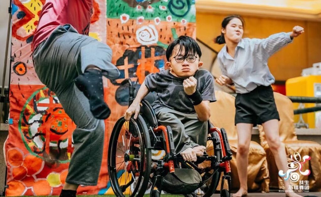 an image of three performers on stage, one is in a wheelchair facing the camera, one on their left is jumping away from camera, and a their performer to their left is dancing. All performers are part of the Chinese performance company Symbiotic for disabled performers.