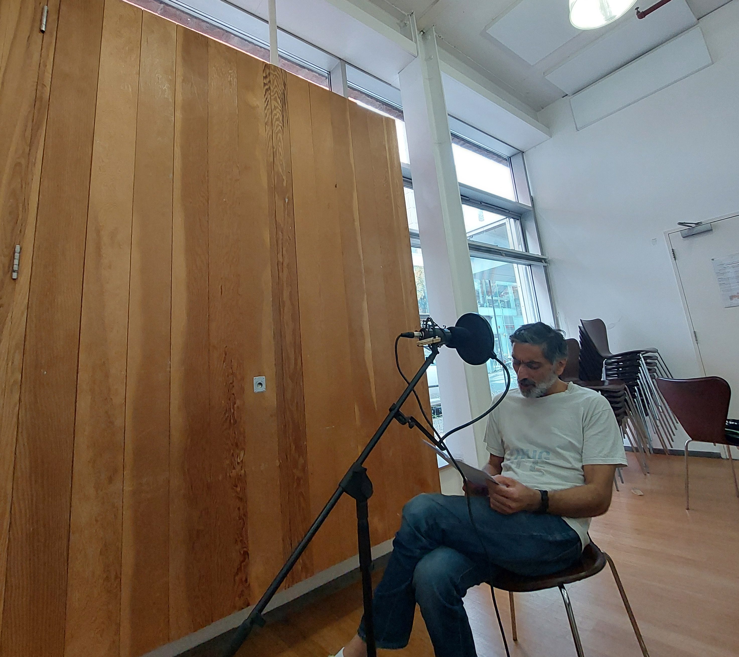 A man is sat on a chair reading a script in front of a microphone. He is in a large room with a wooden wall and large windows behind him.