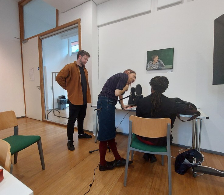 Three people are in a bright room with large windows and wood panneling. One of the sits facing away from the camera recording audio at a desk, the other two are stood near them assisting with their recording.